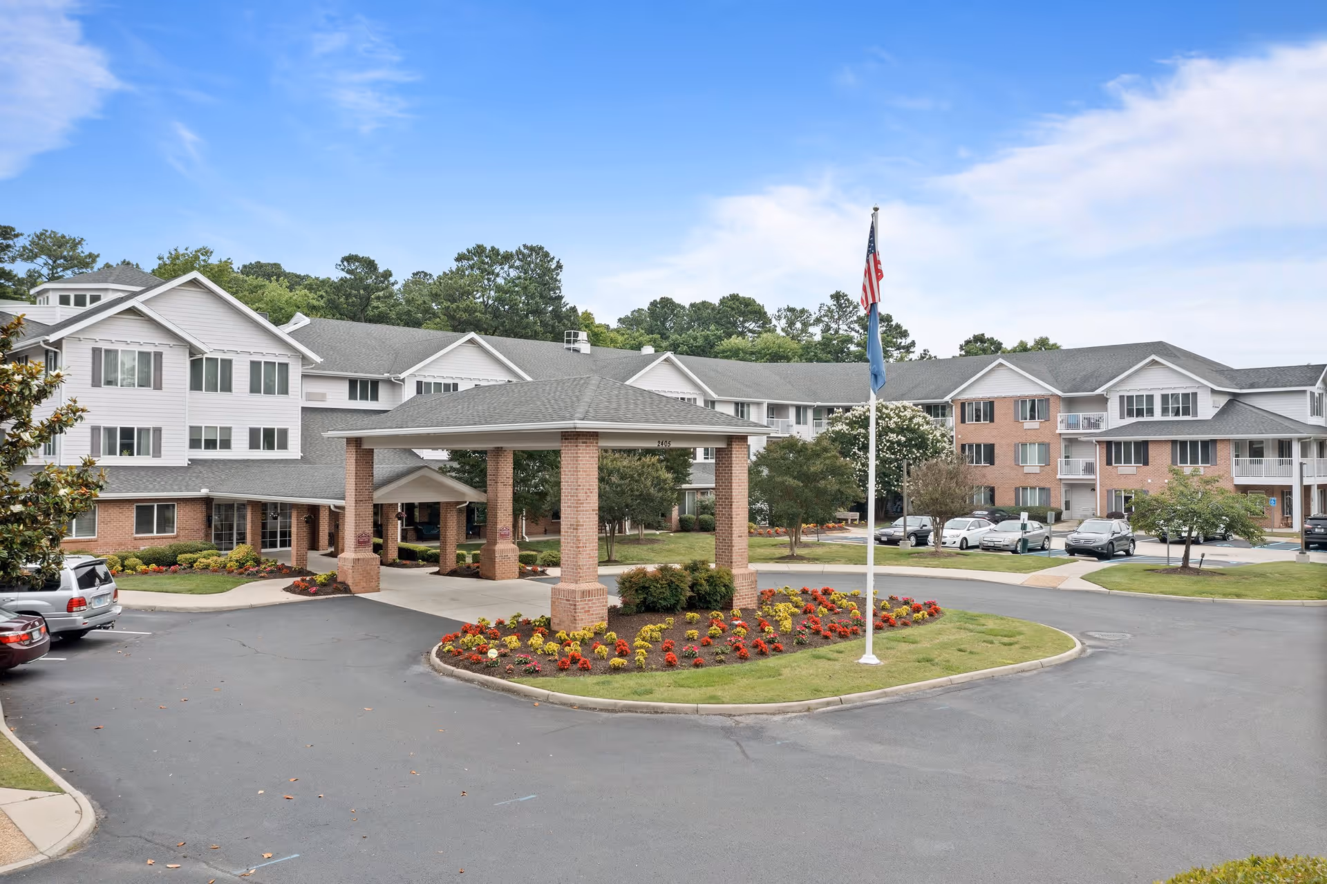 Exterior view of Colonial Harbor Gracious Retirement Living facility showing a large building with multiple windows and a covered entrance supported by brick columns. There is a circular driveway with a landscaped island featuring colorful flowers and a flagpole with the American flag and another flag. Several cars are parked around the building and trees are visible in the background under a partly cloudy sky.