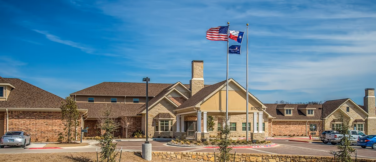 Exterior view of a senior living facility building with a stone and brick facade, three flagpoles displaying the American flag, Texas state flag, and a blue flag, a driveway, and several parked cars under a clear blue sky.