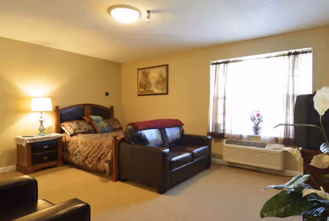Cozy studio bedroom with a bed, leather loveseat, nightstand lamp and a window air conditioner.