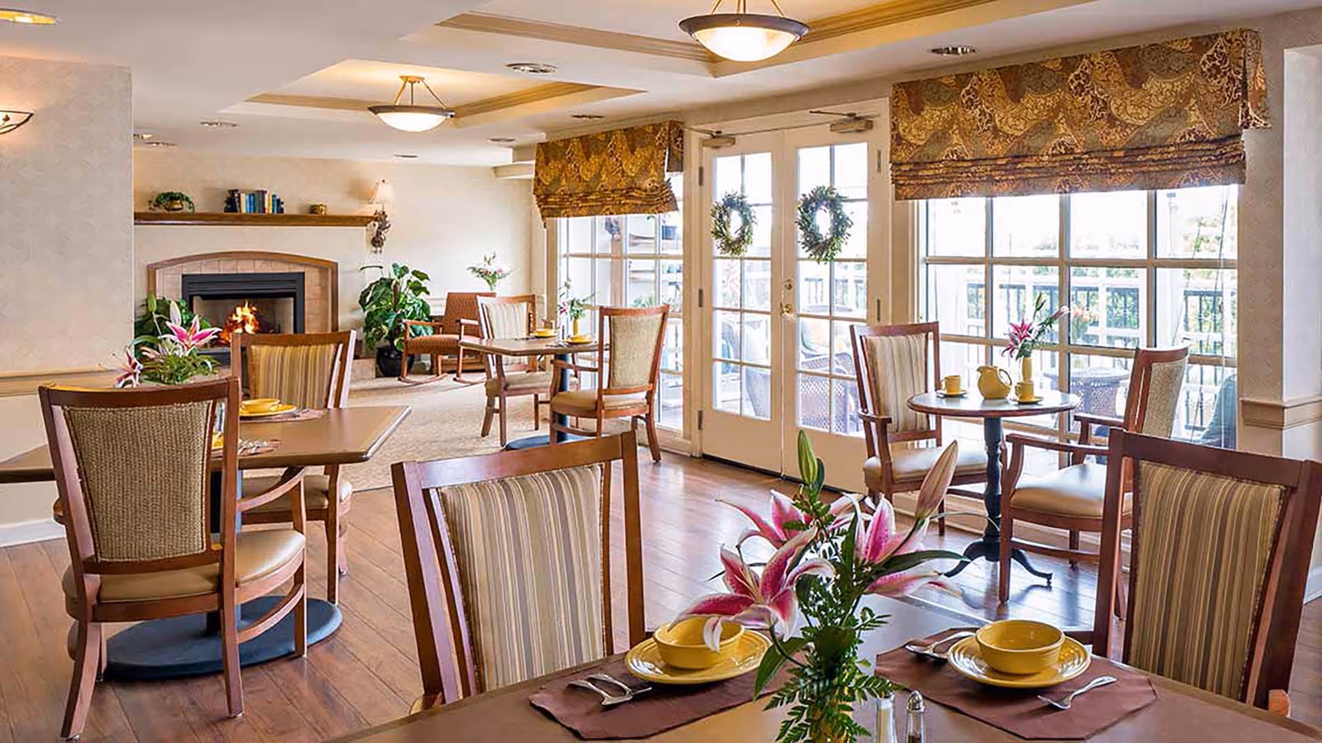 A bright and cozy dining area with wooden tables and chairs, each table set with yellow dishes and floral centerpieces. The room features large windows and glass doors adorned with patterned valances, allowing natural light to fill the space. A fireplace with a mantle holding books and plants is visible in the background, along with additional seating areas.