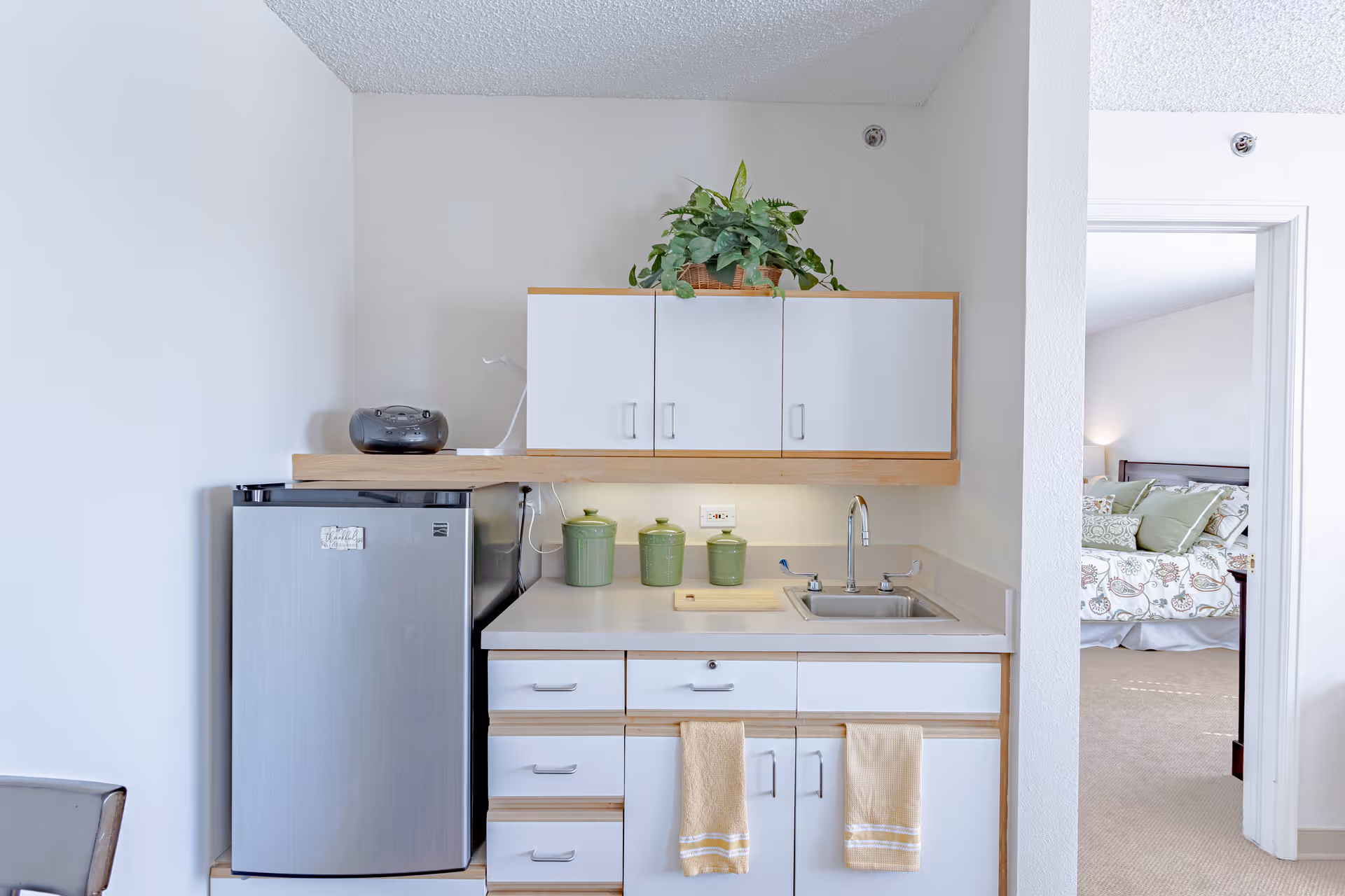 Small kitchenette area with a mini refrigerator, white cabinets, a sink, and a countertop with three green canisters and a cutting board. A potted plant is placed on top of the upper cabinets. To the right, there is an open doorway leading to a bedroom with a bed covered in patterned bedding and multiple pillows.