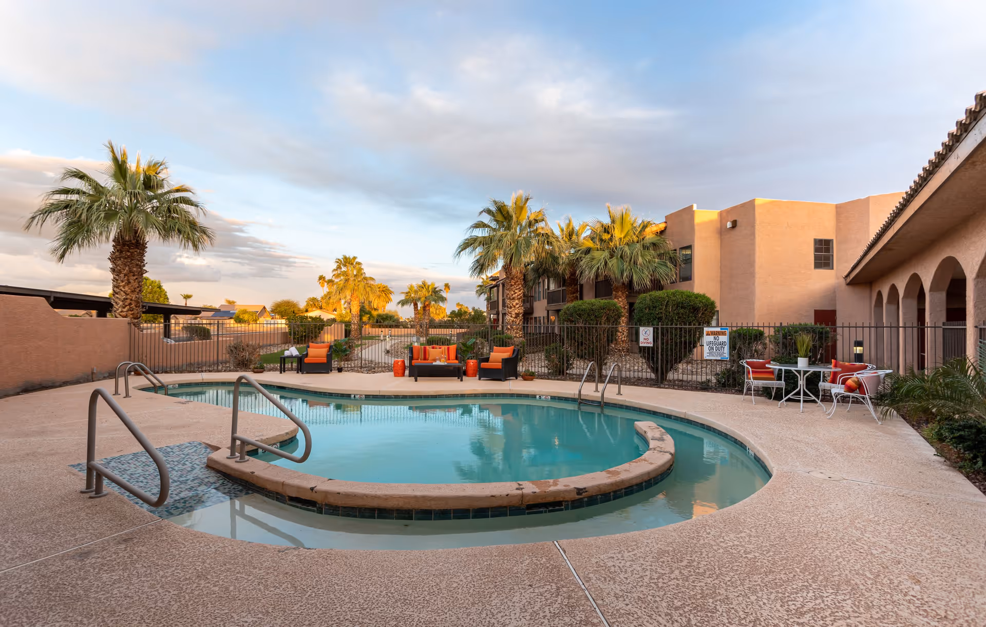 Outdoor swimming pool area at a senior living facility with palm trees, lounge chairs with orange cushions, and a small table with chairs. The building surrounding the pool has a beige exterior and arches along the walkway.