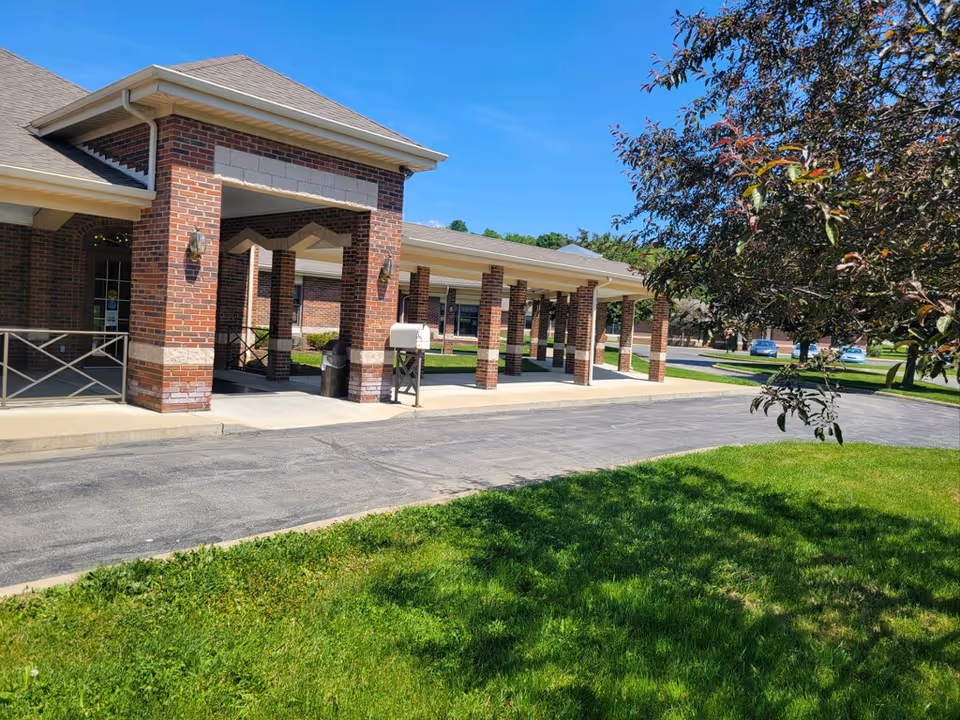 Brick senior care facility entrance with a covered porte-cochere, driveway, and a grassy lawn with a tree.