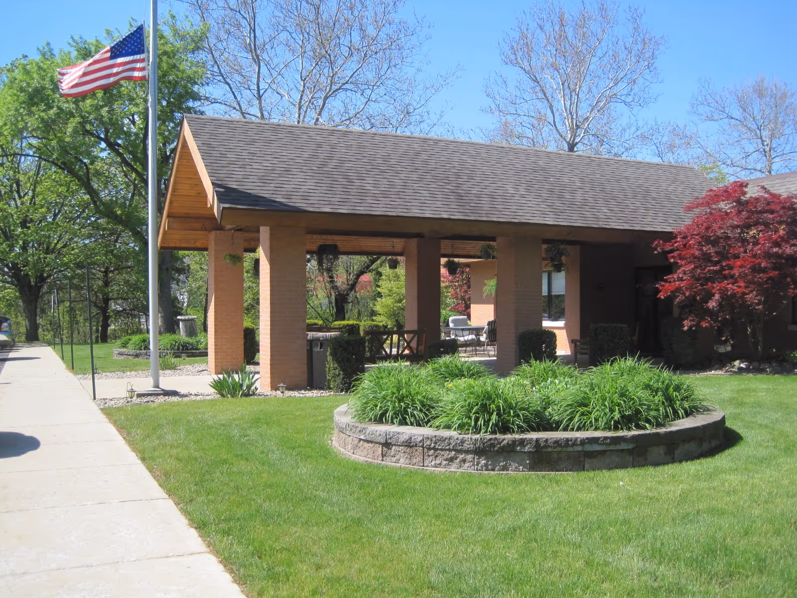 Covered brick building entrance with a flagpole, circular raised planter and green lawn under a clear blue sky.
