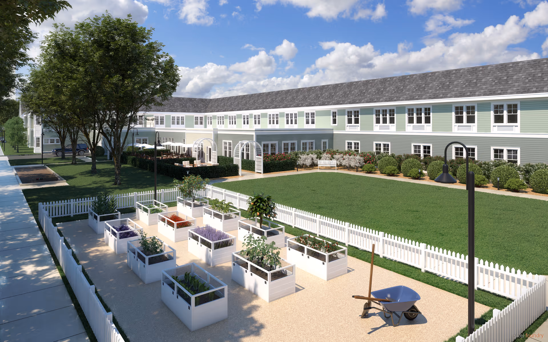 Outdoor garden area at a senior living facility with raised garden beds containing various plants, a wheelbarrow, white picket fences, green lawn, trees, and a two-story building with multiple windows in the background under a partly cloudy sky.