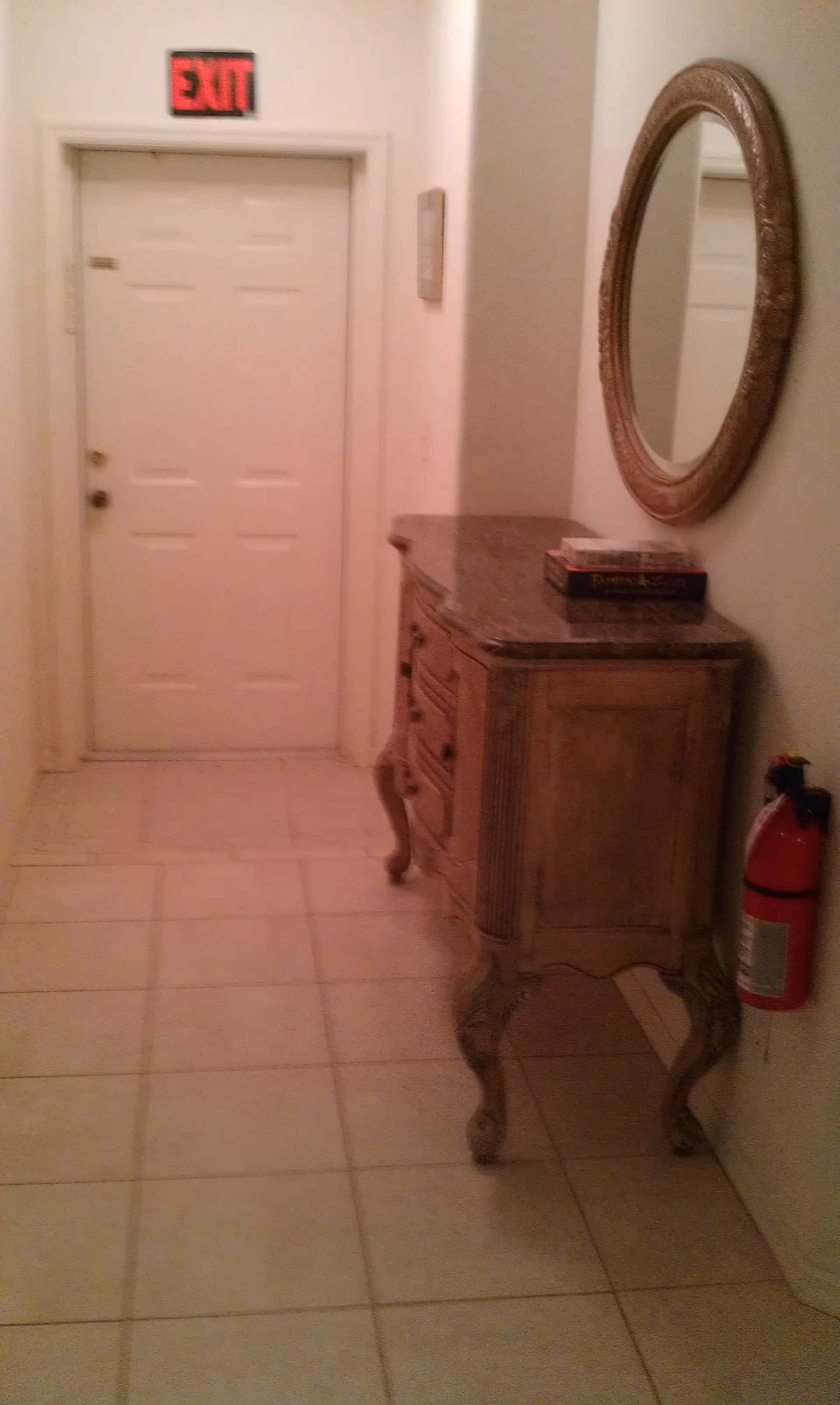 A narrow hallway with tiled floor leading to a white door with an illuminated red EXIT sign above it. On the right side of the hallway, there is an ornate wooden console table with a marble top, holding two stacked books. Above the table is a round decorative mirror with a carved frame. A red fire extinguisher is mounted on the wall near the table.