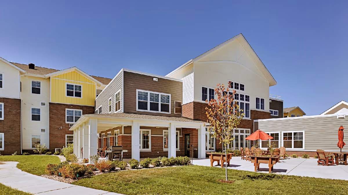 Courtyard patio with wooden tables, umbrellas, and chairs in front of a multi-story senior living building under a clear blue sky.