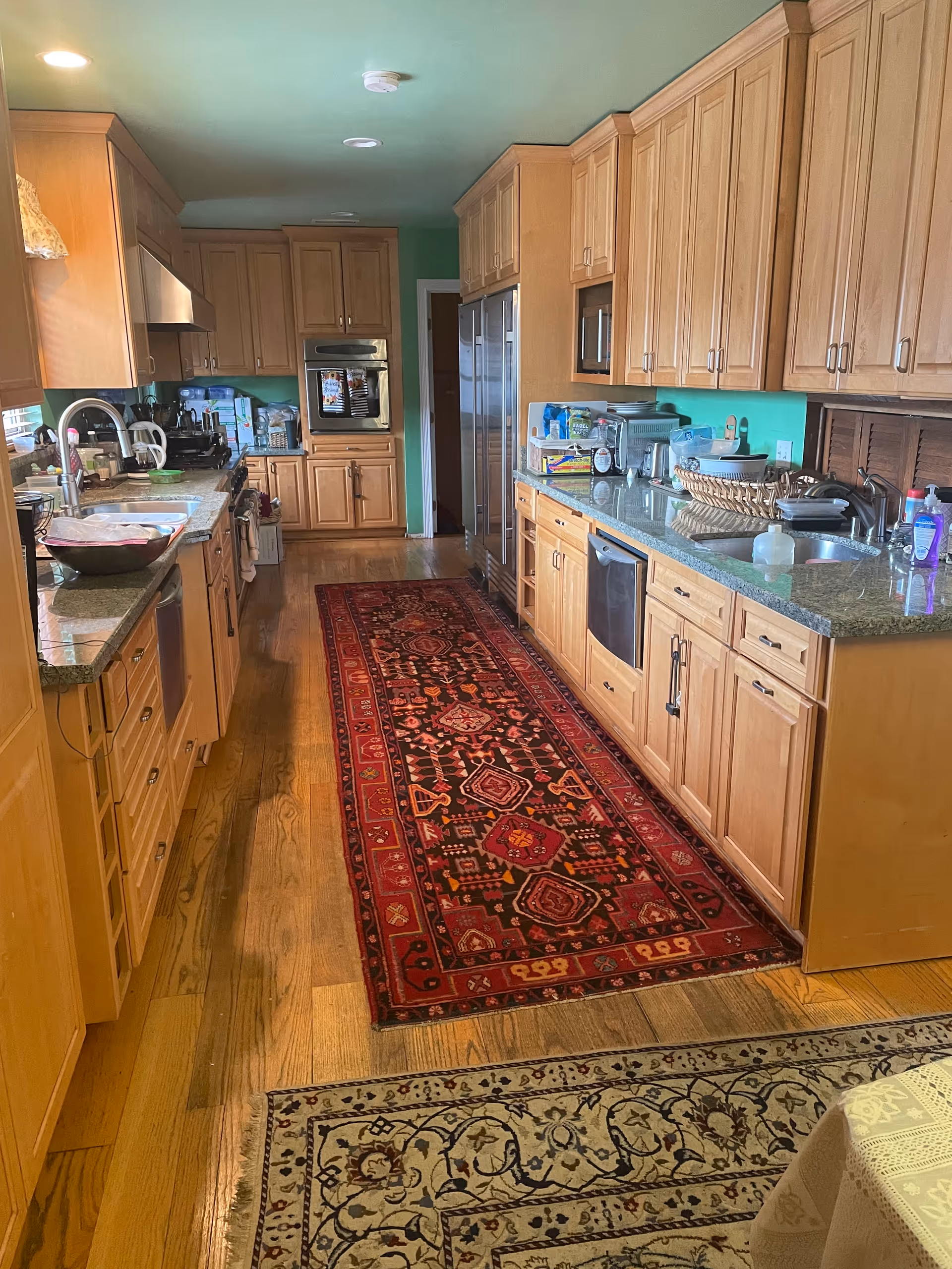 Galley kitchen with wooden cabinets, granite countertops, stainless appliances and a long red runner rug down the center.