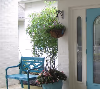 A covered outdoor porch area with a blue bench, a hanging basket with plants, a large potted plant with flowers and small American flags, and a light blue door with glass panels.