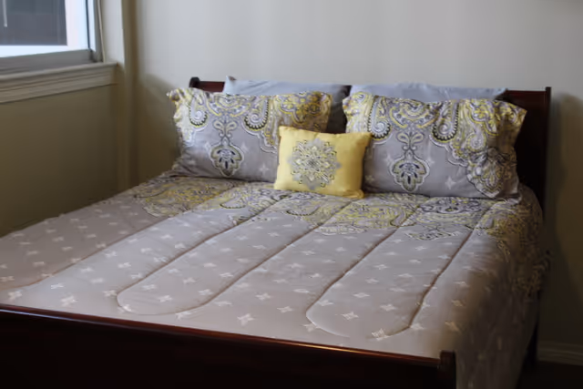 A neatly made bed with a wooden frame, adorned with a comforter and pillows featuring a gray and yellow paisley pattern. There is a small yellow decorative pillow in the center. A window is visible to the left of the bed.