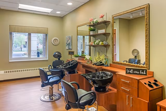 Interior of a hair salon area in a senior living facility with two black salon chairs in front of two black hair washing sinks. There are large mirrors on the wall above wooden cabinets and shelves decorated with plants and flowers. A window with a beige valance lets in natural light, and a clock and wall art are visible on the wall.
