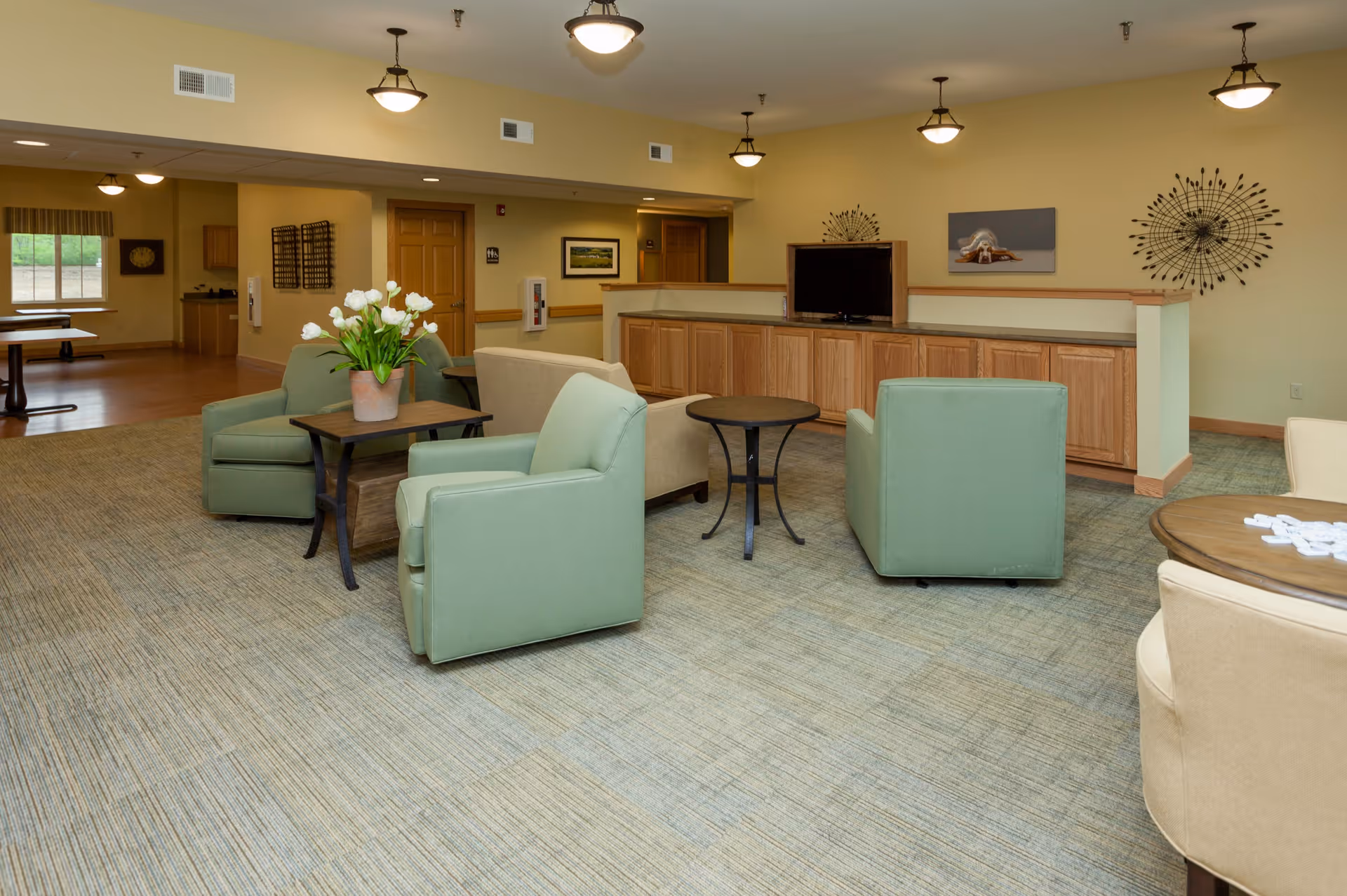 A spacious senior living facility common area with green and beige armchairs arranged around small round tables. A TV is placed on a wooden cabinet against the wall, which is decorated with modern metal wall art and a framed picture. The room has beige walls, carpeted flooring, and ceiling lights. In the background, there is a dining area with tables and chairs near windows.