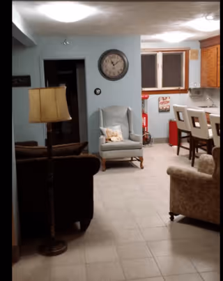 Interior view of a living area with a light blue armchair, a floor lamp, a brown sofa, and a beige armchair. The room has tiled flooring and light blue walls. A clock is mounted on the wall above the armchair. In the background, there is a kitchen area with wooden cabinets, a countertop with bar stools, and a window.