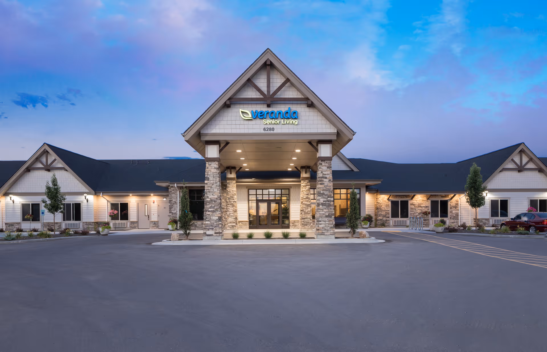 Front exterior view of a senior living facility building named Veranda Senior Living, featuring a large entrance with stone pillars, a peaked roof, and well-lit surroundings during dusk with a clear sky.