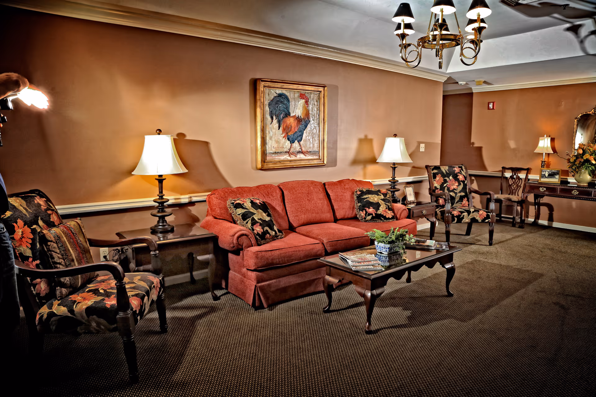 A cozy living room with a red sofa, two floral-patterned armchairs, a wooden coffee table with magazines and a plant, two side tables with lamps, a chandelier, and a painting of a rooster on the wall.