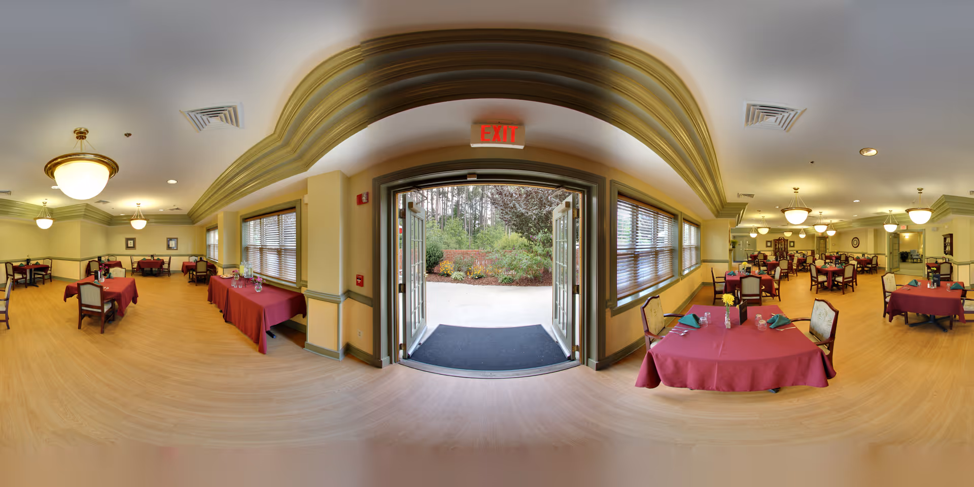 Interior view of a dining room in a senior living community with multiple tables covered in red tablecloths, set with plates, glasses, and napkins. The room features large windows with blinds, warm lighting from ceiling fixtures, and an open double door leading outside to a garden area.