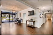 Interior view of a senior living facility lobby with wooden flooring, two dark chairs near a large window, a wall-mounted flat screen TV above a small table with a plant, and a hallway leading to other rooms.