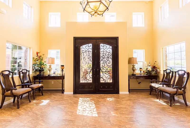 Bright and spacious entryway with large double doors featuring intricate wrought iron designs. The room has high ceilings with multiple windows allowing natural light to fill the space. There are two sets of wooden chairs with cushioned seats on either side, accompanied by small tables adorned with lamps and floral arrangements.