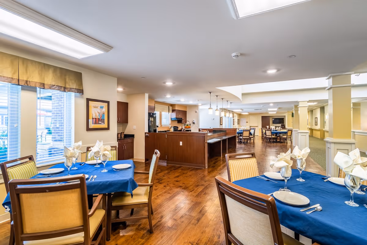 A spacious dining area in a senior living facility with tables covered in blue tablecloths, set with plates, glasses, and folded napkins. The room features wooden floors, large windows with yellow valances, and a kitchen area with dark wood cabinetry in the background.