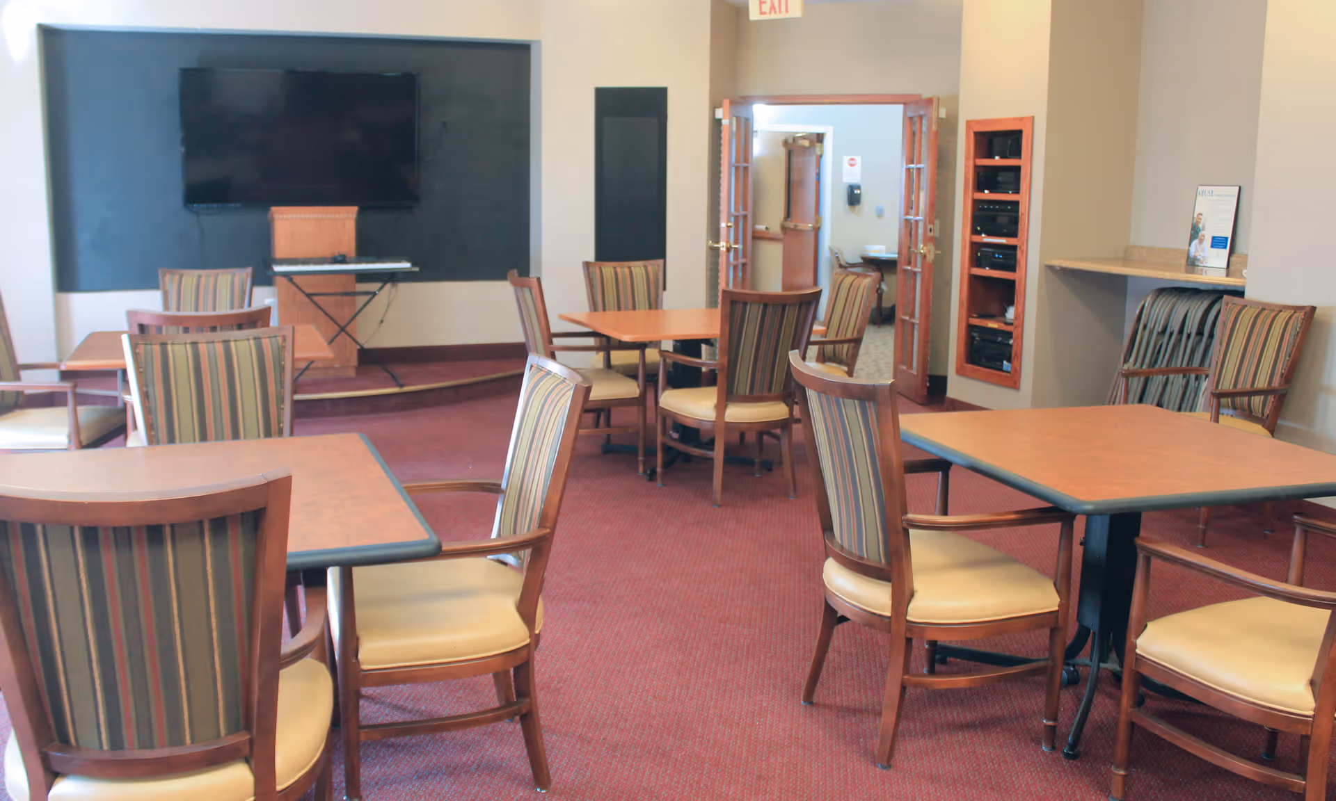A common area with several wooden tables and chairs with striped upholstery on a red carpet. A large flat-screen TV is mounted on a black wall panel, with a keyboard on a stand below it. There are double doors leading to another room, and a rack with audio-visual equipment is mounted on the wall to the right.