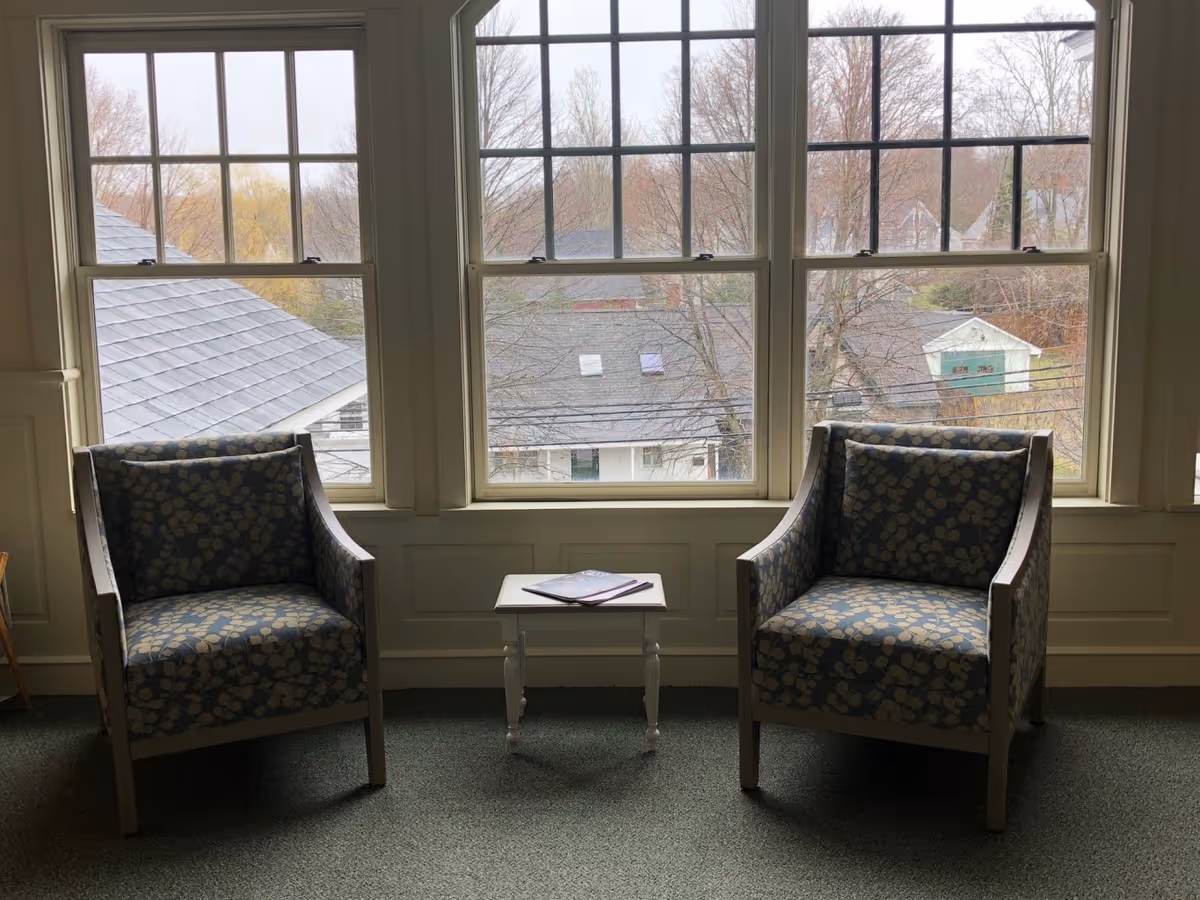Two patterned armchairs with a small white table between them, placed in front of large windows showing a view of rooftops and trees outside.