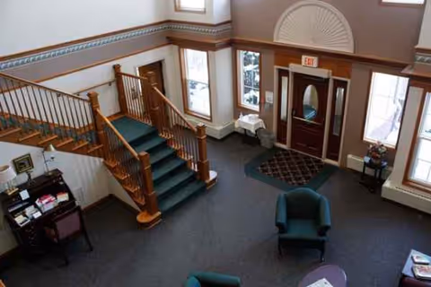 Senior living facility lobby with a wooden staircase, seating, and double entrance doors.