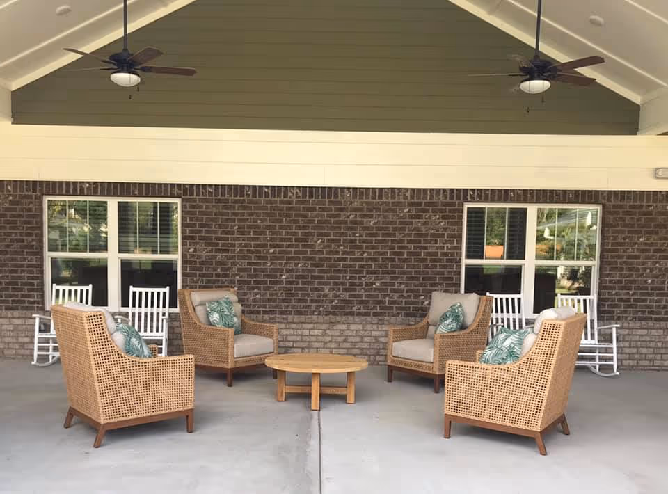 Covered outdoor seating area with four wicker armchairs around a low round wooden table in front of a brick wall and windows, with rocking chairs and ceiling fans overhead.