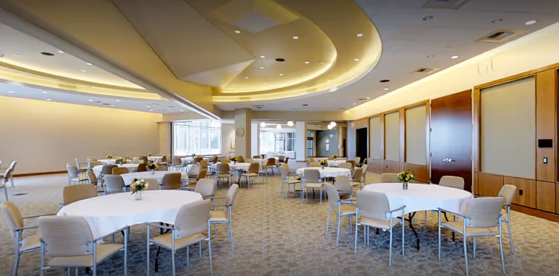 A spacious dining room with multiple round tables covered with white tablecloths, each surrounded by beige chairs. Small floral centerpieces are placed on each table. The room features a patterned carpet, large windows letting in natural light, and a ceiling with recessed lighting and a curved design.