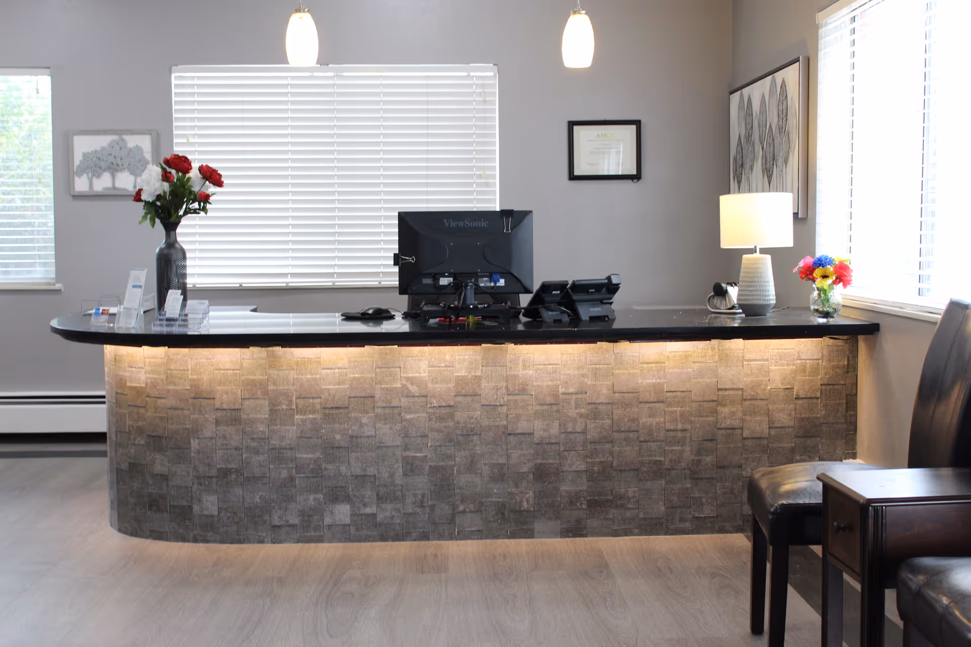 Reception desk area with a black countertop and textured front panel illuminated by under-counter lighting. On the desk are a computer monitor, two telephones, a vase with red and white flowers, a lamp, and a small bouquet of colorful flowers. Behind the desk are two windows with white blinds, a framed certificate, and a framed artwork. To the right are two black chairs and a small wooden table.