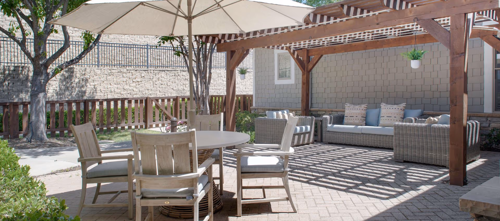 Outdoor patio area with a round table and four wooden chairs under a large umbrella. There is a wooden pergola with hanging plants and cushioned wicker sofas with pillows underneath. The area is paved with bricks and surrounded by a wooden fence and trees.