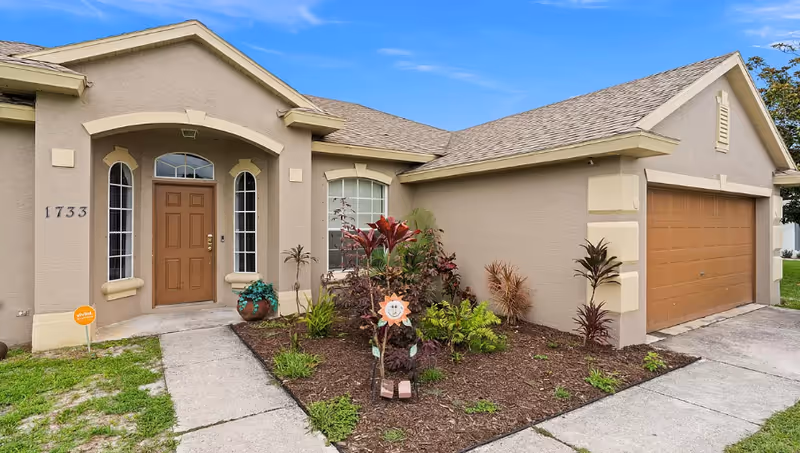 Front exterior of a beige single-story house with a brown front door, attached garage, and landscaped flowerbed by the walkway.