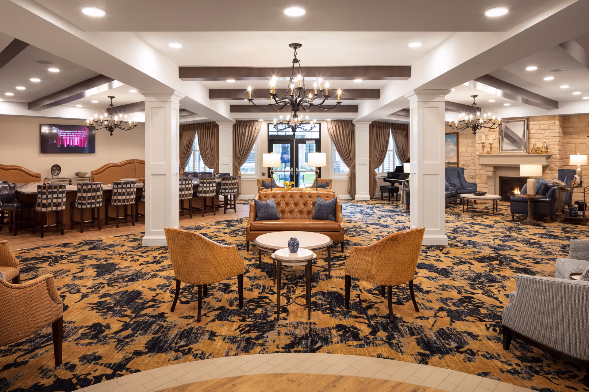 Spacious, elegantly furnished senior living lounge with multiple seating areas, chandeliers, a fireplace, and a dining/bar area in the background.