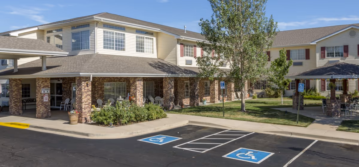 Exterior view of Crossroads Senior Living at Northglenn showing a two-story building with beige siding and brick pillars. There is a covered entrance with chairs, a small garden area, several trees, and two handicap parking spaces in front. The sky is clear and blue.