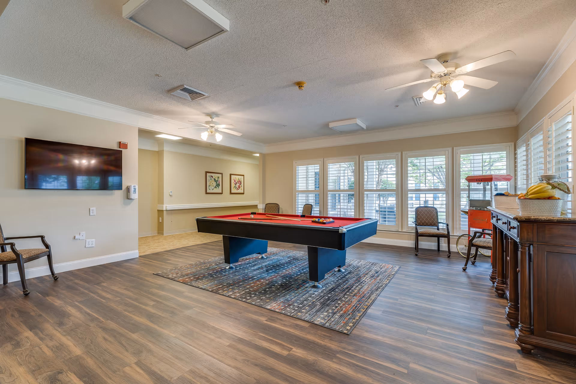 A spacious recreational room with a red felt pool table on a patterned rug in the center. The room has large windows with white shutters, several chairs, a wall-mounted flat screen TV, ceiling fans with lights, and a wooden sideboard with a basket of bananas and a popcorn machine.