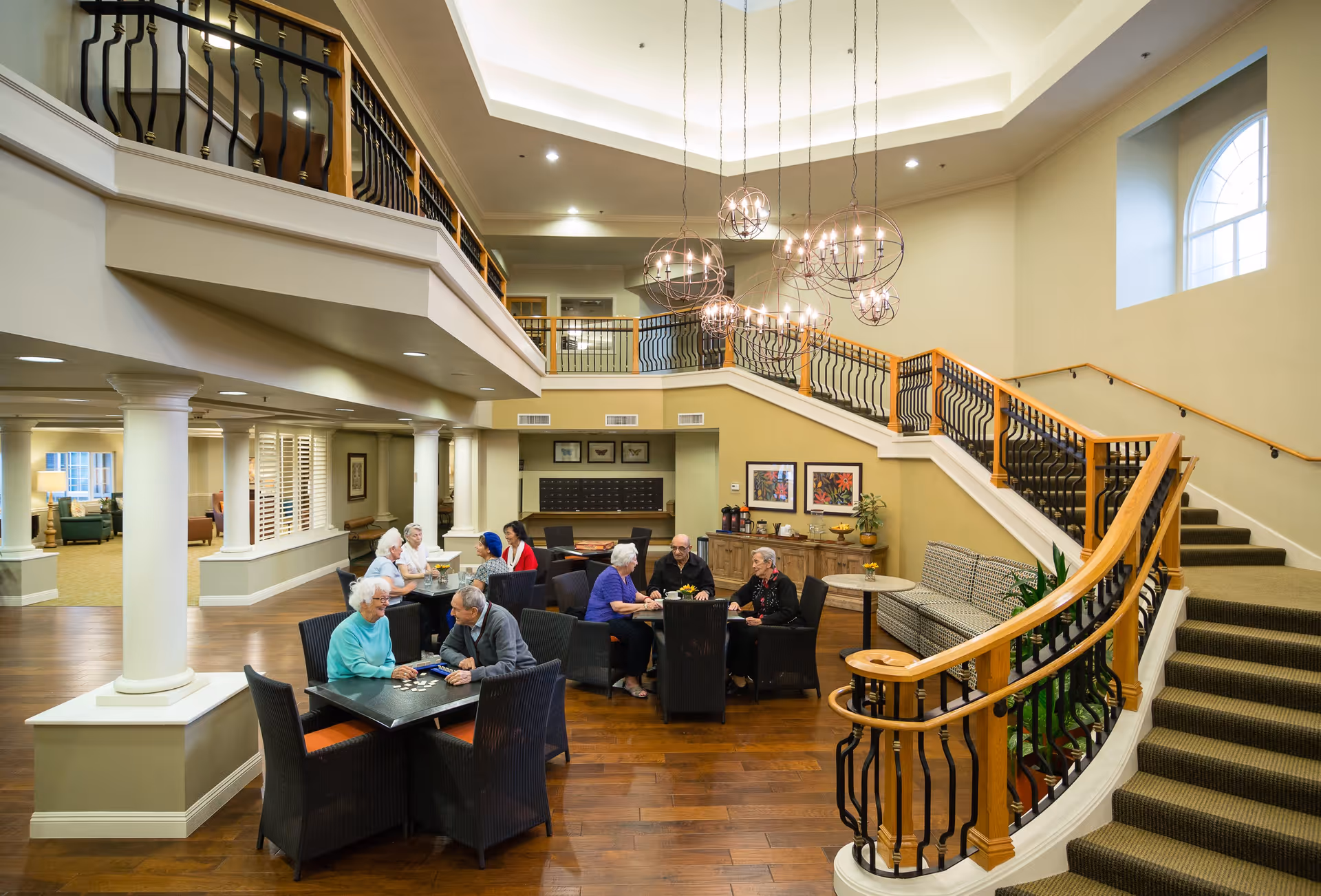A spacious and well-lit common area in a senior living facility with several elderly people sitting at tables engaged in conversation and activities. The room features a wooden floor, a staircase with wooden handrails and black balusters, modern hanging light fixtures, and large windows allowing natural light to enter.