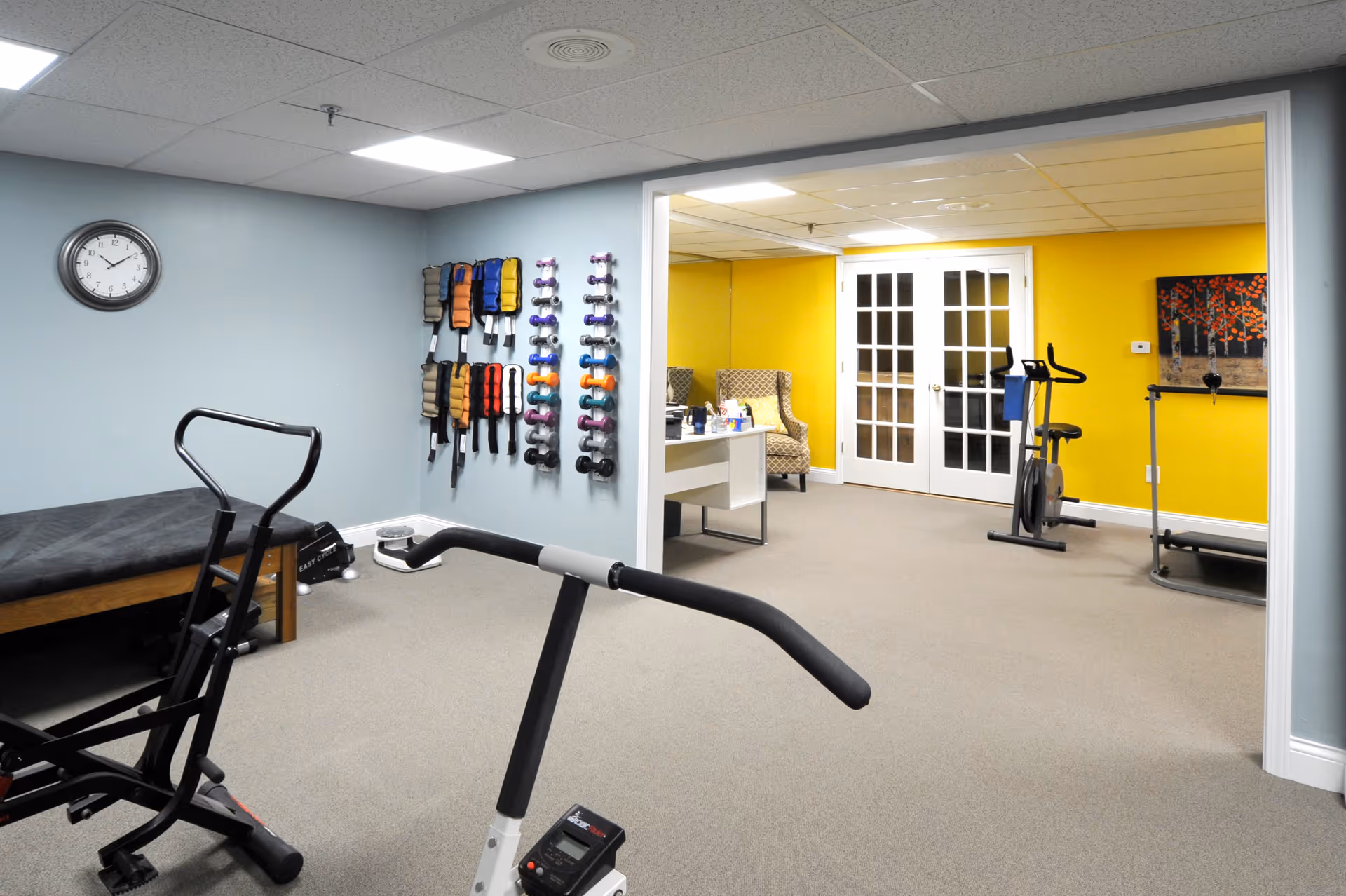 A fitness room with exercise equipment including a stationary bike, weights on wall racks, and a treadmill. The room has light blue and yellow walls, a clock on the wall, and a small seating area with two armchairs and a table in the background.