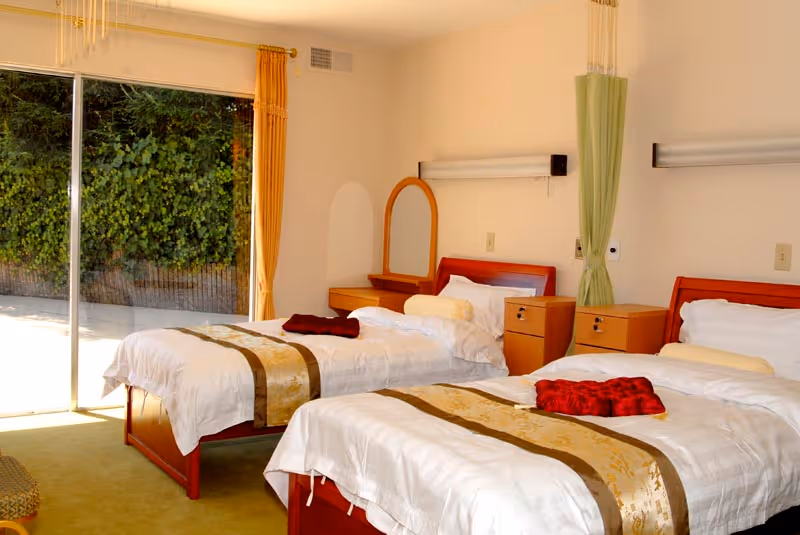 A bright room with two single beds neatly made with white linens and gold-accented runners. Each bed has a red folded blanket on top. Between the beds are two wooden nightstands with drawers. A wooden vanity with a mirror is positioned against the wall. A large sliding glass door on the left side opens to an outdoor area with greenery visible outside. The room has light-colored walls and carpeted floor.