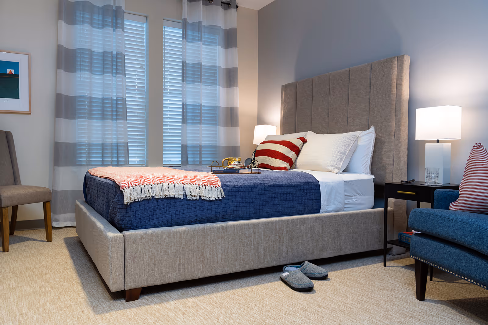 A neatly made bed with a beige upholstered headboard and frame, covered with a blue quilt and white sheets, accented by a red and white striped pillow and a pink throw blanket. Next to the bed is a black nightstand with a white lamp and a glass of water. A pair of gray slippers is placed on the beige carpet floor. The room has two windows with white blinds and gray and white striped curtains. A blue armchair with a red and white striped pillow and a beige chair are also visible.