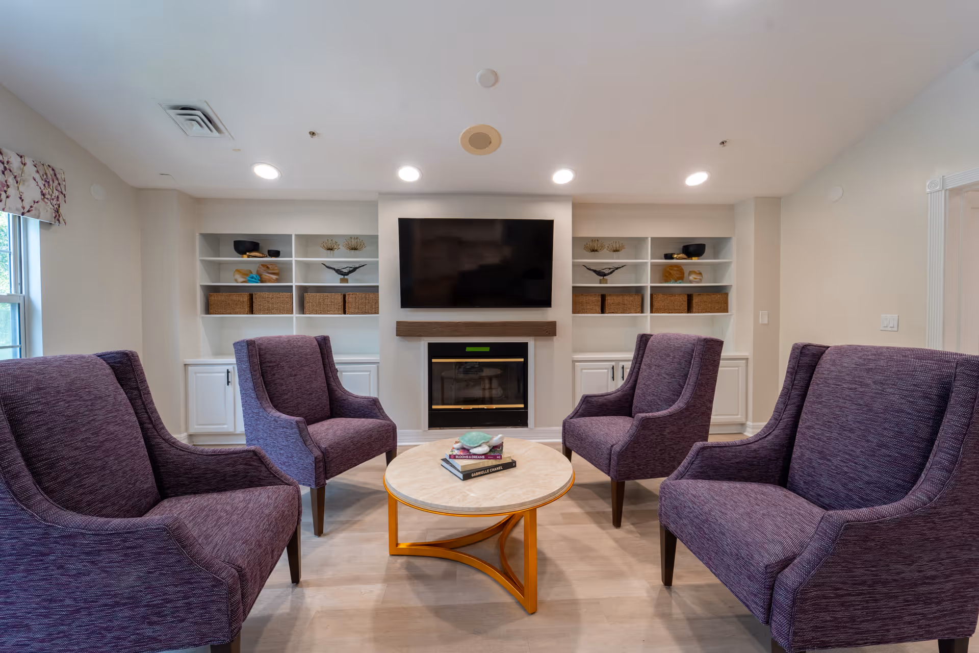 A cozy living room with four purple upholstered armchairs arranged around a round wooden coffee table with books and a decorative item on it. The room features built-in white shelves with baskets and decorative pieces, a fireplace, and a large flat-screen TV mounted above the fireplace. The walls are light-colored and the room is well-lit with recessed ceiling lights.