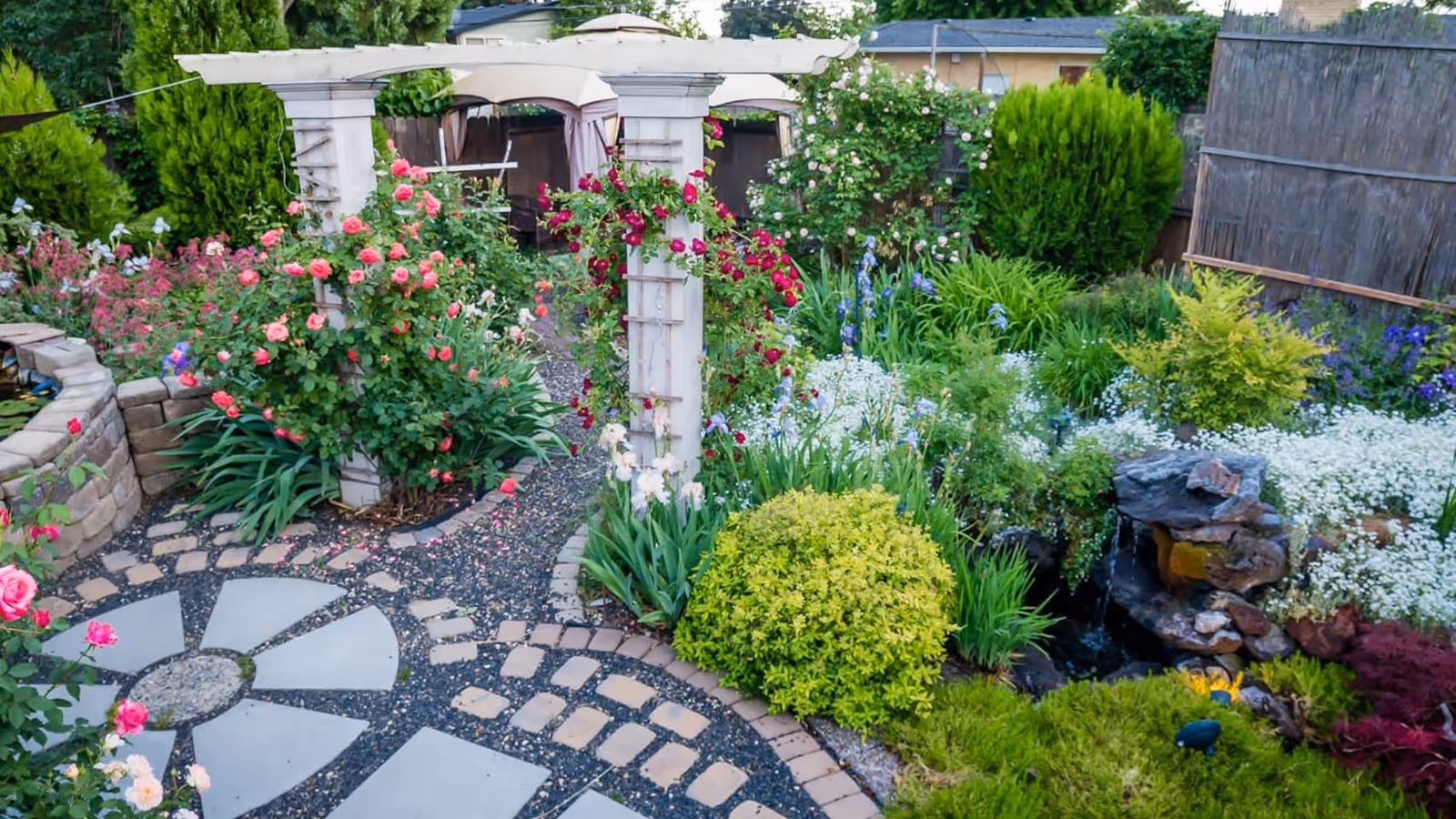 A beautifully landscaped garden with a stone pathway, colorful flowers including roses, green shrubs, and a small rock water feature. There is a white wooden pergola with climbing roses and a gazebo with seating in the background.
