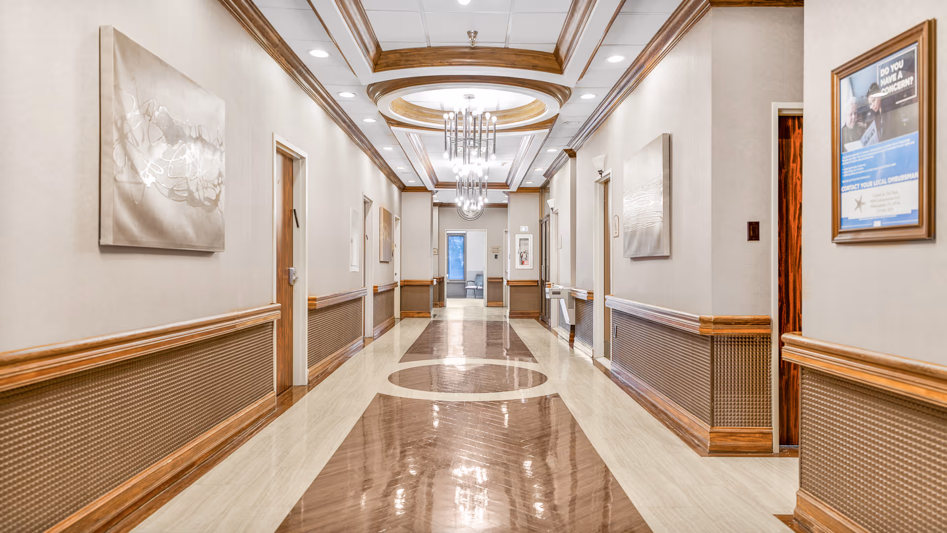 A long, well-lit hallway in a healthcare facility with polished floors featuring circular and rectangular patterns. The walls are beige with wooden trim and wainscoting. Several doors line both sides of the hallway, and framed artwork and informational posters are hung on the walls. Modern chandeliers hang from the ceiling, and a water fountain is visible on the right side.