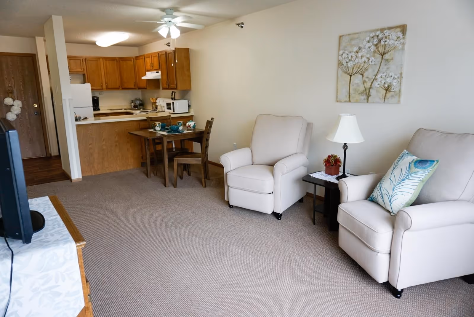 A cozy living area with two beige armchairs, a small side table with a lamp and a small plant, a wooden dining table set with dishes, and a kitchen with wooden cabinets and white appliances in the background.