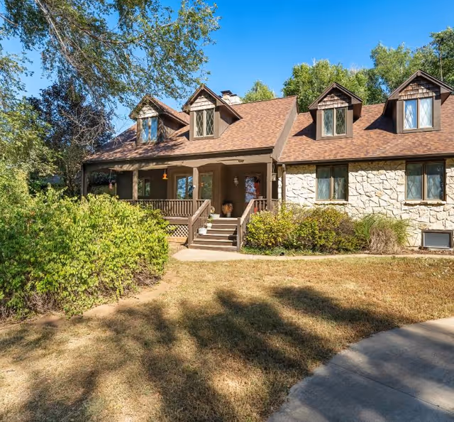 Front view of a stone-faced house with a covered porch, dormer windows, and a grassy yard.
