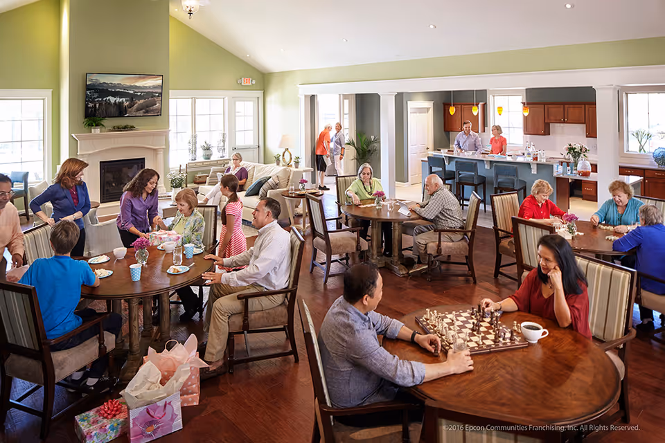 A lively senior living community common area with multiple round wooden tables where elderly residents and visitors are engaged in various activities such as playing chess, eating, and socializing. The room has large windows letting in natural light, a fireplace with a mounted TV above it, and an open kitchen area with bar stools in the background. The space is warmly decorated with plants and flowers, and the atmosphere appears friendly and welcoming.