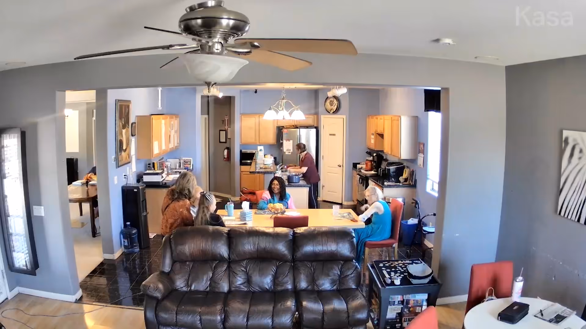 Open living room and kitchen with a leather sofa in the foreground and several residents seated around a table.
