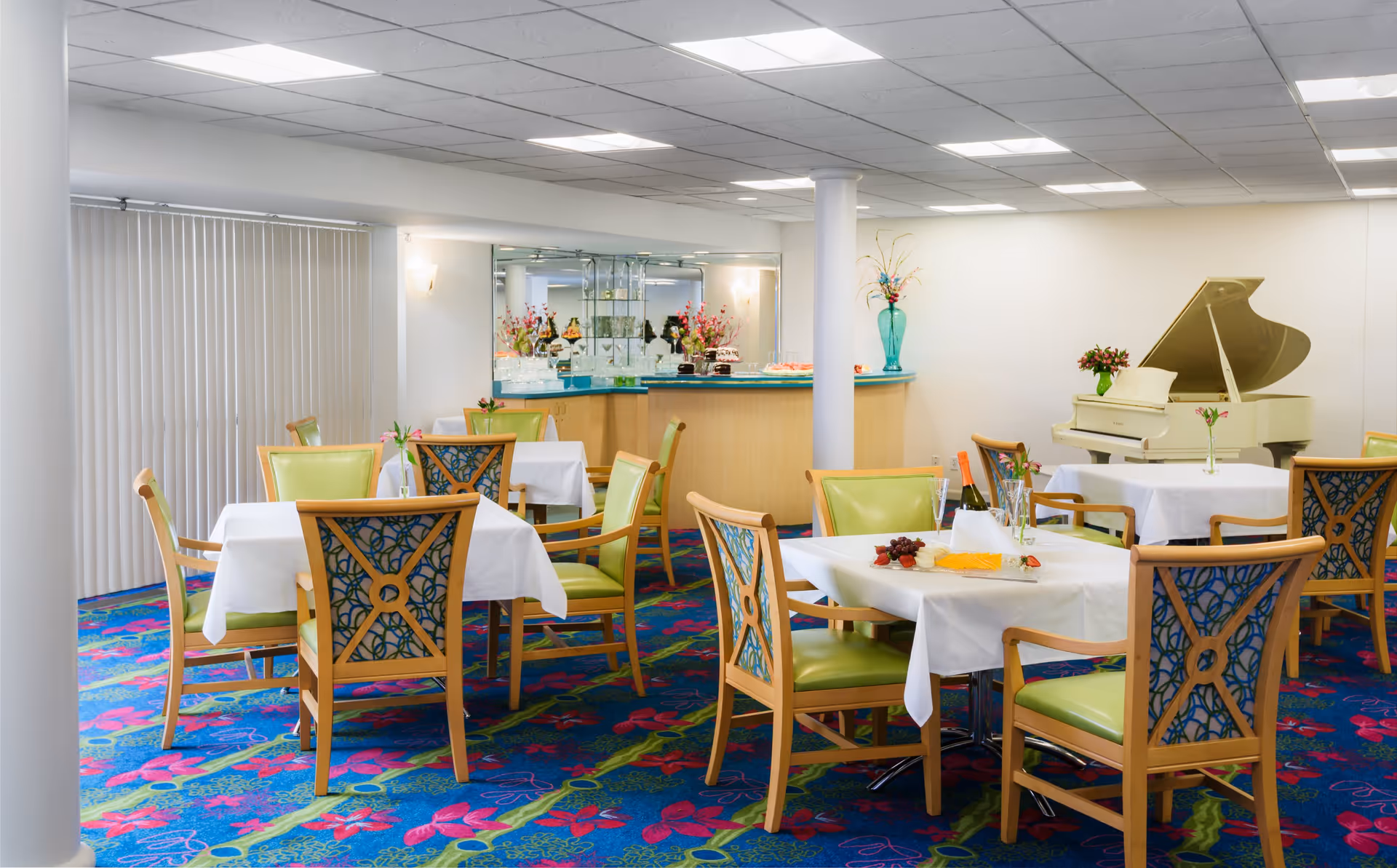 A bright dining room with several tables covered in white tablecloths, surrounded by wooden chairs with green cushions and decorative backs. The room features a colorful carpet with a floral pattern, a white grand piano in the corner, and a small bar area with glass shelves and floral arrangements. Vertical blinds cover the windows on one side of the room.
