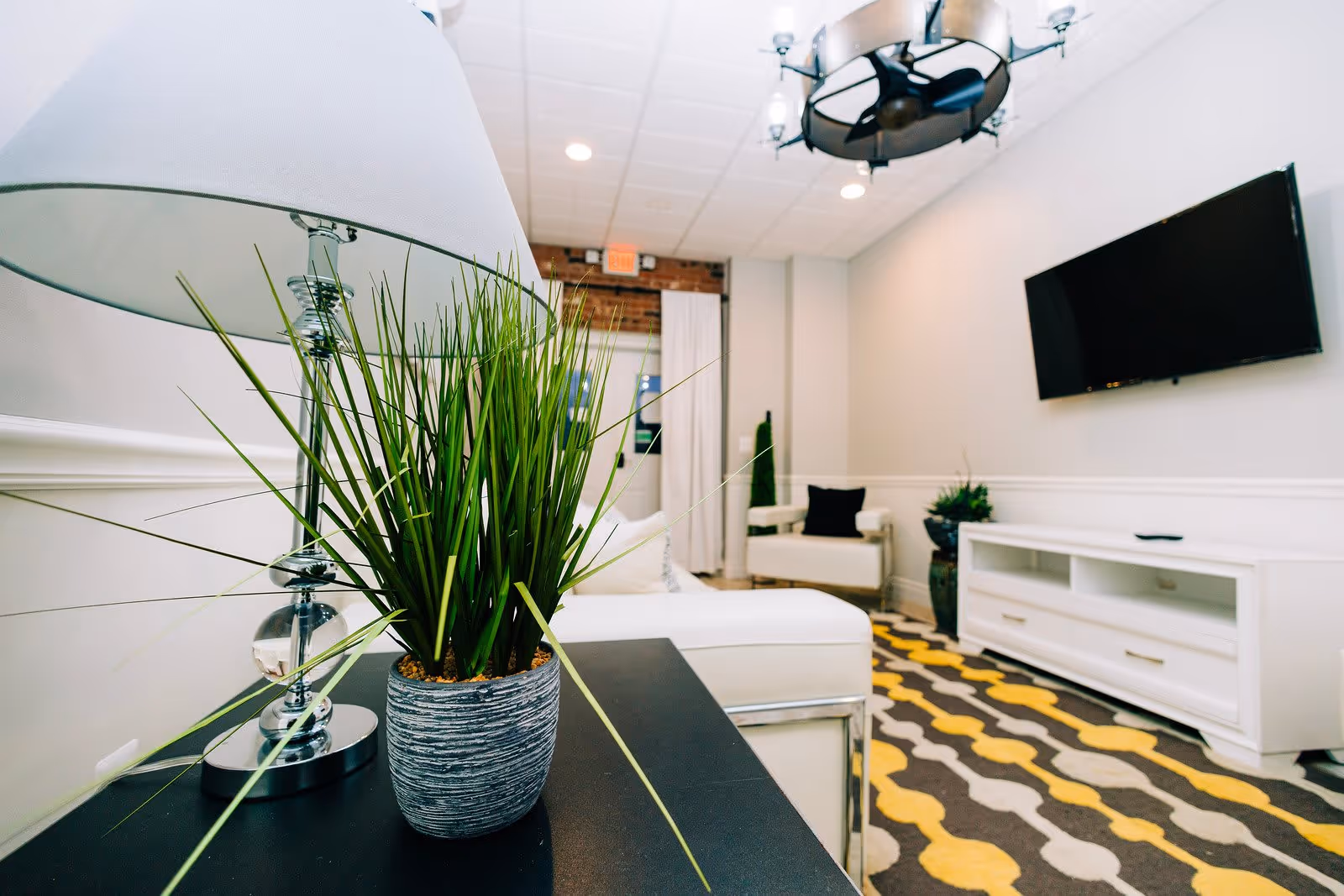 A modern living room with a white couch, a white armchair, a black and yellow patterned rug, a flat-screen TV mounted on the wall, a white TV stand, a decorative green plant in a gray pot on a black table, and a silver lamp with a white lampshade. The ceiling has recessed lighting and a circular chandelier.