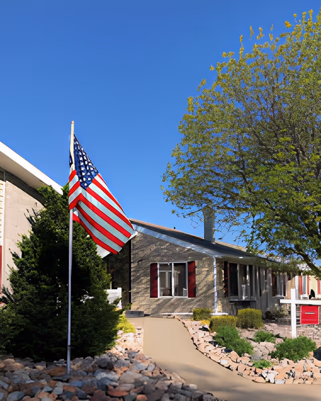Exterior view of Lebensraum Assisted Living facility showing a single-story building with red window shutters, a large tree to the right, an American flag on a flagpole to the left, and a paved walkway leading to the entrance surrounded by landscaping and rocks.