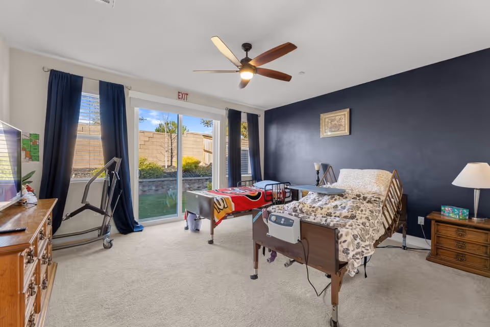 Bright care facility bedroom with two adjustable hospital-style beds, sliding glass doors to a small yard, dark accent wall, and bedside furniture.