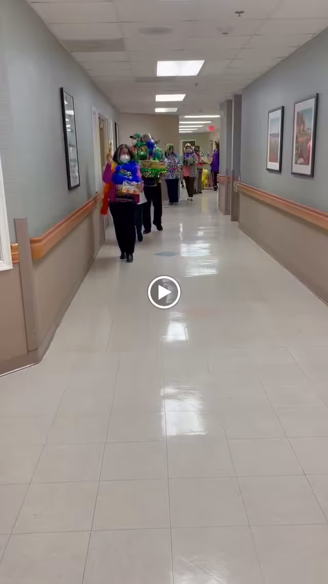 A group of people walking down a long, clean hallway in a senior living facility. The hallway has beige walls with wooden handrails on both sides and framed pictures hanging on the right wall. The people are wearing colorful clothing and some are holding items, with one person in front wearing a face mask.